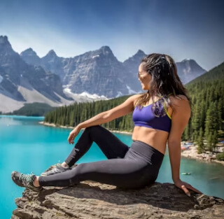 Young woman wearing exercise clothing, sitting on the edge of a cliff overlooking aqua blue water and a mountain landscape.