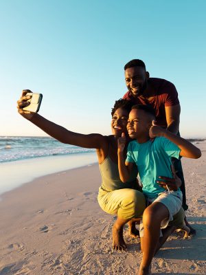 African american woman taking selfie with cheerful husband and son over smartphone at beach. Copy space, sunset, unaltered, family, together, sky, childhood, nature, vacation, enjoyment, technology.